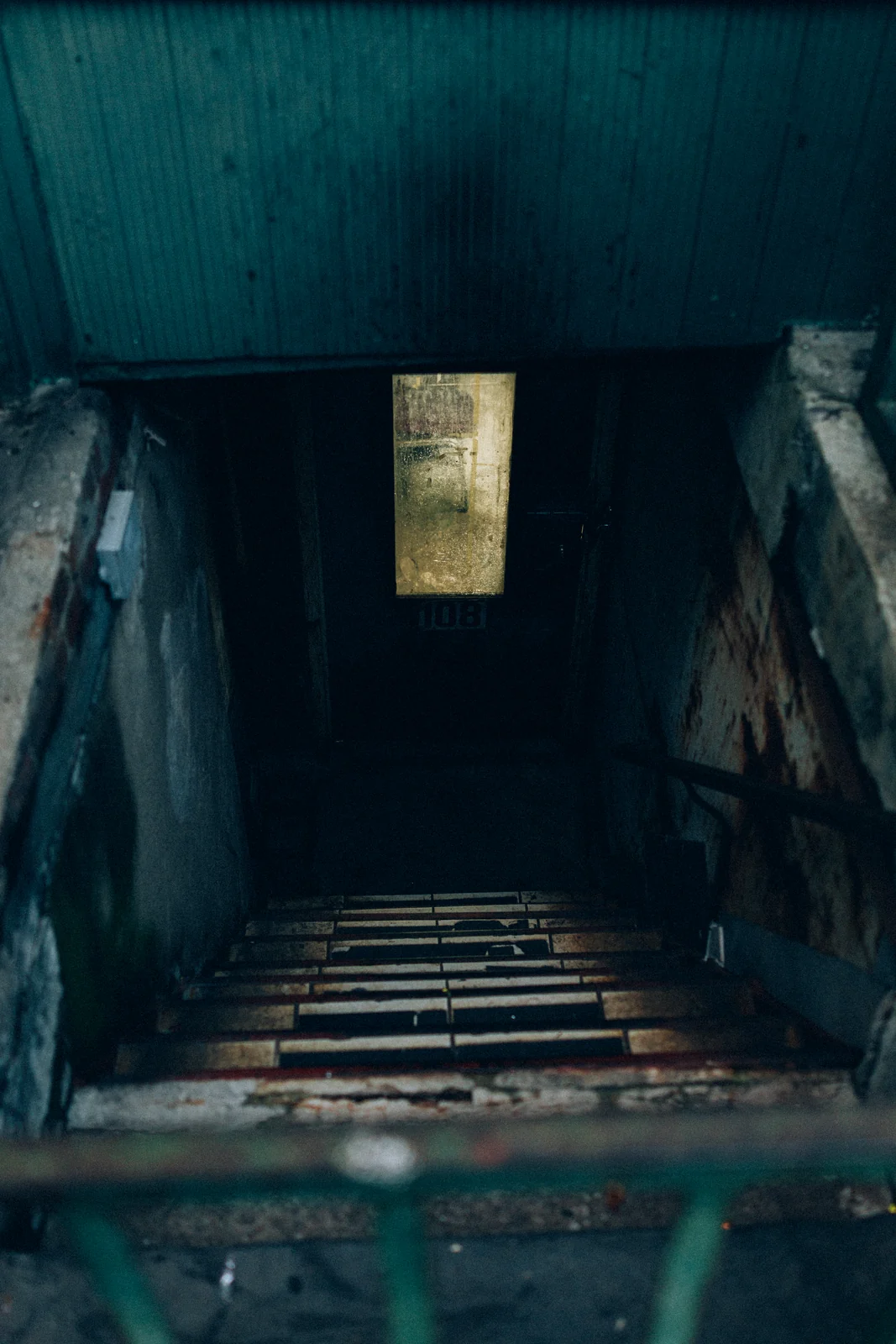 A steel-railed stairwell descending to a lit basement doorway, graded with GX-05 108