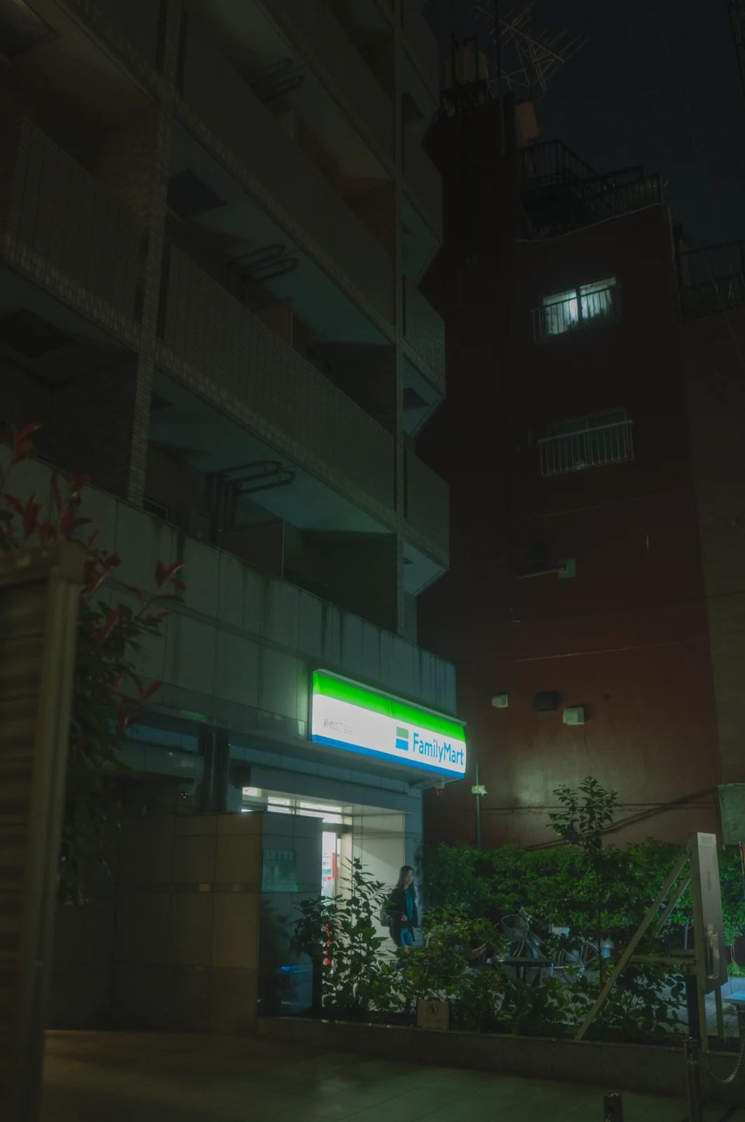 A lit convenience store at the foot of a dark apartment block, graded with GX-06 Family Mart