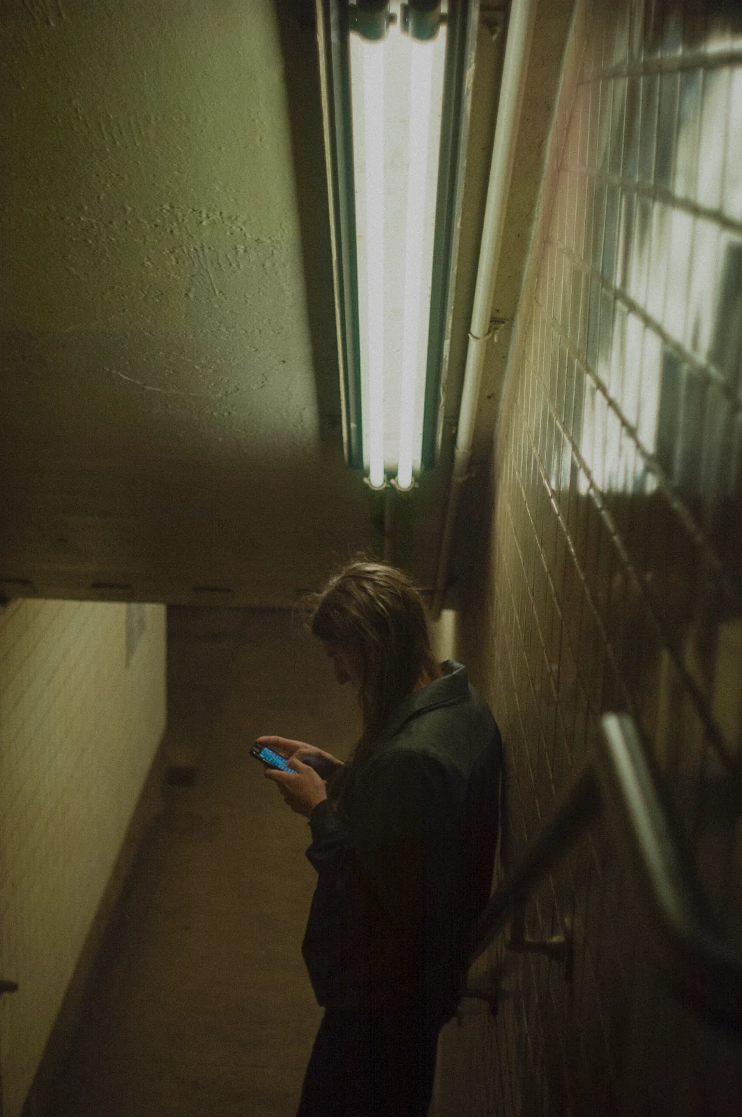 A figure on a phone in a fluorescent-lit stairwell, graded with GX-06 Fluorescent Underground