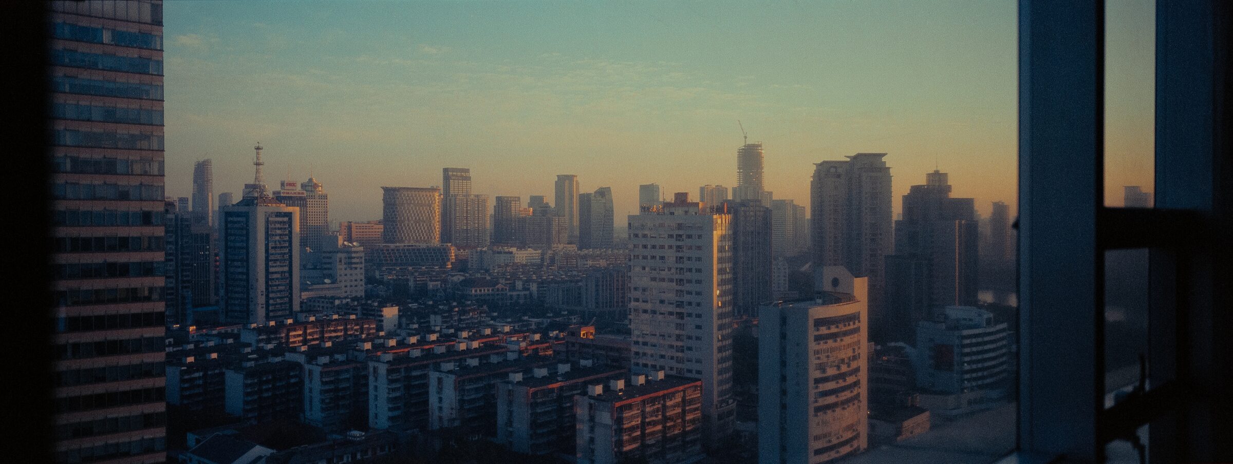 City skyline at dusk seen past a high window frame, warm haze over dense apartment blocks
