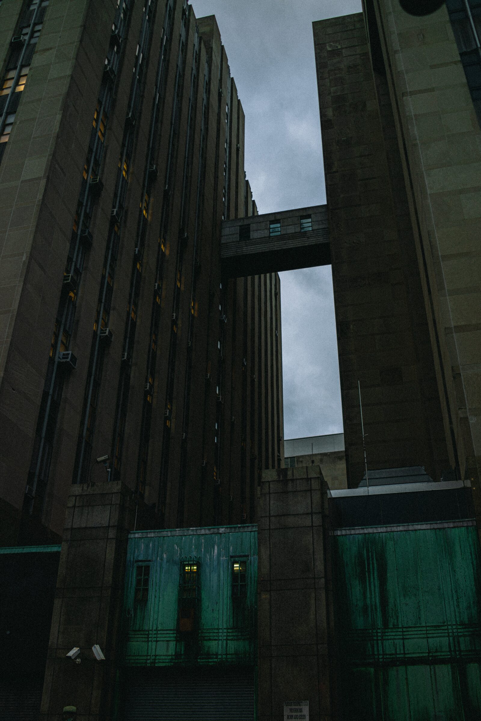 A brutalist concrete tower and skybridge above weathered green shutters under overcast sky
