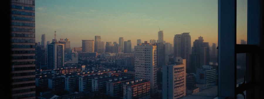 City skyline at dusk seen past a high window frame, warm haze over dense apartment blocks