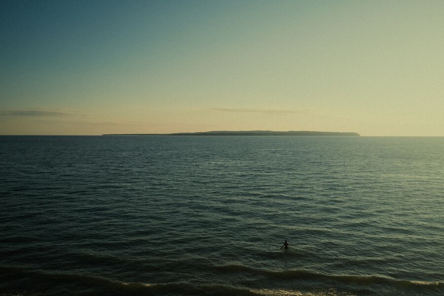 A lone surfer on open water at dusk with a distant headland under a pale sky
